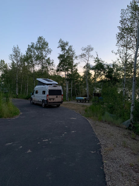 An RV rental camper van with a pop-top is parked on a paved road at a wooded campsite at dusk.