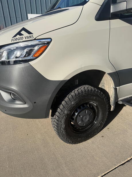 Front-side view of a white Forged Vans Sprinter camper van, equipped with Outpost A/T tires. Excellent for RV rental adventures.