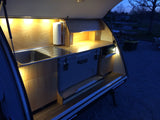 Compact rv rental camper kitchen, illuminated with warm lights. Features a sink, cooler, and organized storage for camping.