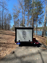 This compact RV rental, with a dreamcatcher decal, is perfectly parked by a lake, offering a serene outdoor escape.