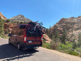 Adventure-ready RV rental with bikes. A red camper van is parked on a scenic road, surrounded by majestic layered mountains.