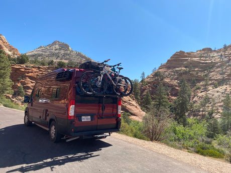 Adventure-ready RV rental with bikes. A red camper van is parked on a scenic road, surrounded by majestic layered mountains.