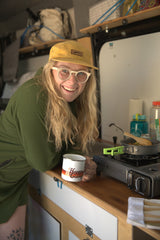 Blonde woman in an RV rental smiles, holding a "Happy Camp" mug while cooking on a portable stove. Perfect for van life!
