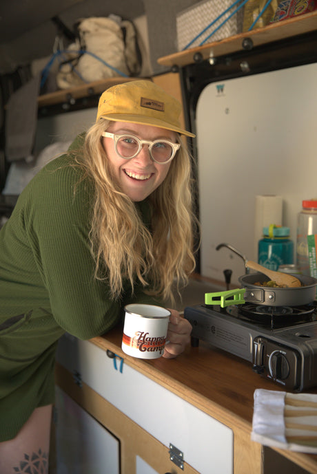 Blonde woman in an RV rental smiles, holding a "Happy Camp" mug while cooking on a portable stove. Perfect for van life!