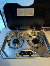 Clean two-burner stainless steel gas cooktop in an RV rental, with the lid open to reveal lighting instructions.