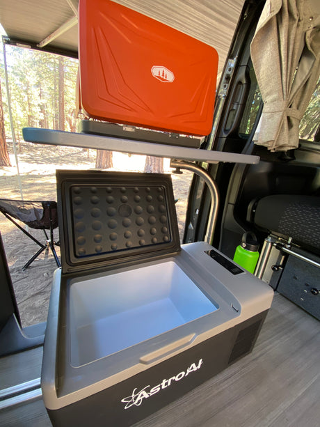 RV rental: Portable fridge with open lid, orange camping stove on a shelf, and van interior looking out to a forest.