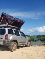 Silver Nissan Xterra, rooftop tent on sandy beach by ocean. Explore overlanding, a rugged alternative to an rv rental.
