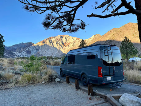 Scenic mountain views from a grey Sprinter camper van. This RV rental is parked at a dirt campsite under a clear blue sky, framed by pine branches.