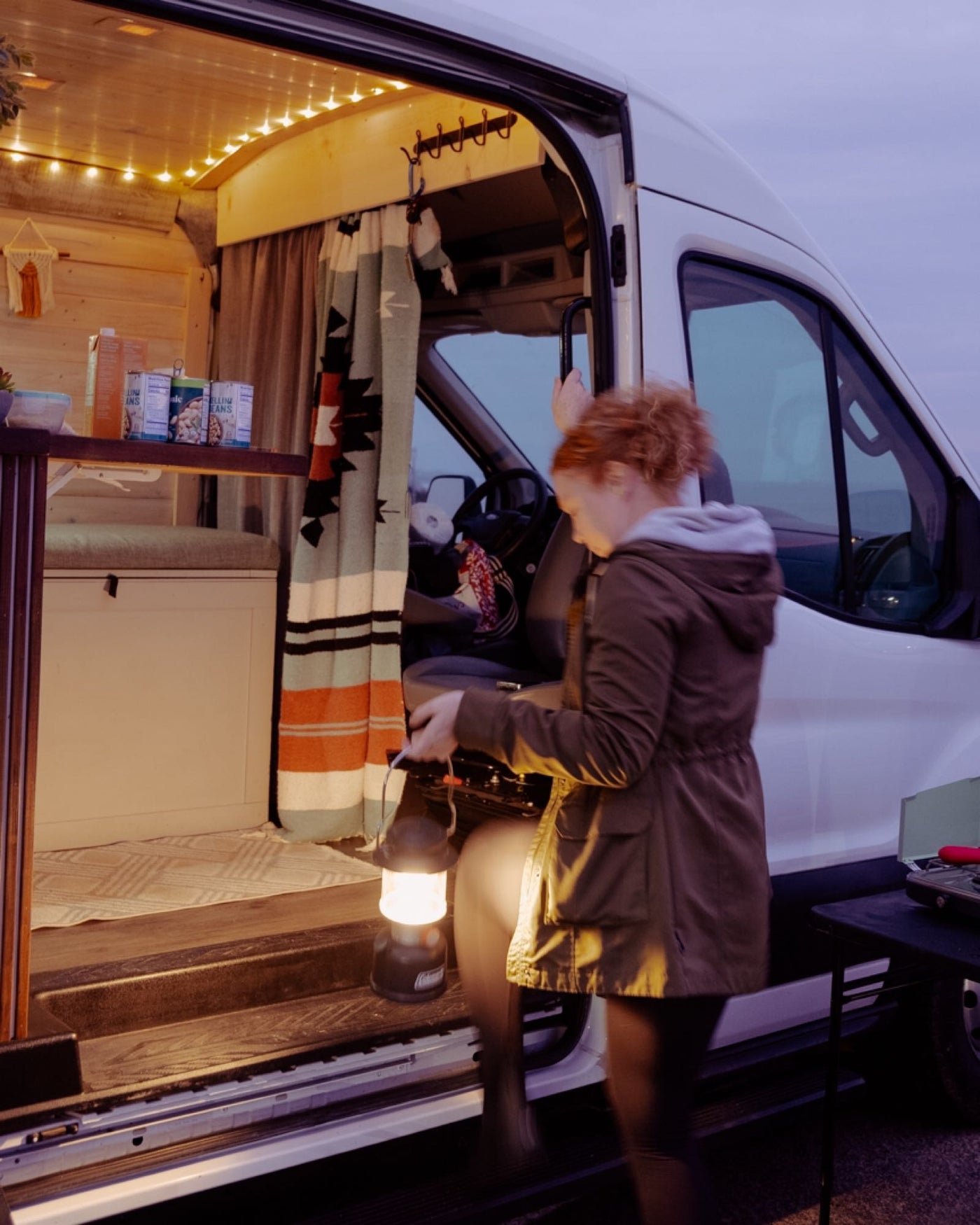 Red-haired person exits a cozy RV rental van with a lantern. Interior string lights illuminate the evening. — photo 10