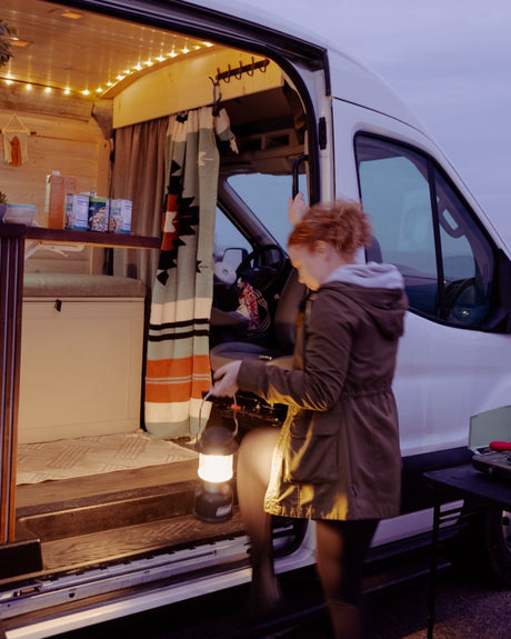 Red-haired person exits a cozy RV rental van with a lantern. Interior string lights illuminate the evening.