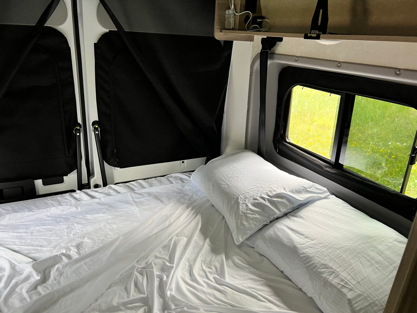 Inside a comfy RV rental: A neat bed with white sheets and pillows next to windows showing a bright green outdoor scene. Ideal for travel rest.