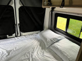 Inside a comfy RV rental: A neat bed with white sheets and pillows next to windows showing a bright green outdoor scene. Ideal for travel rest.