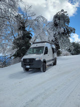 This white Mercedes Sprinter RV rental is on a snowy road, surrounded by stunning ice-covered trees.