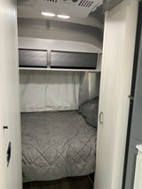 Inside a modern RV rental, a compact bedroom features a gray quilted bed, white privacy curtains, and overhead storage.