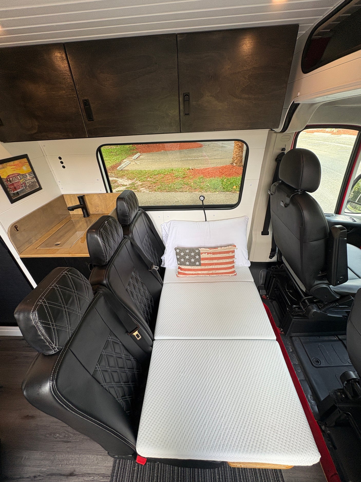 An RV rental interior showcasing a comfortable foldable bed, black leather seats, overhead cabinets, and a compact kitchen sink area.
