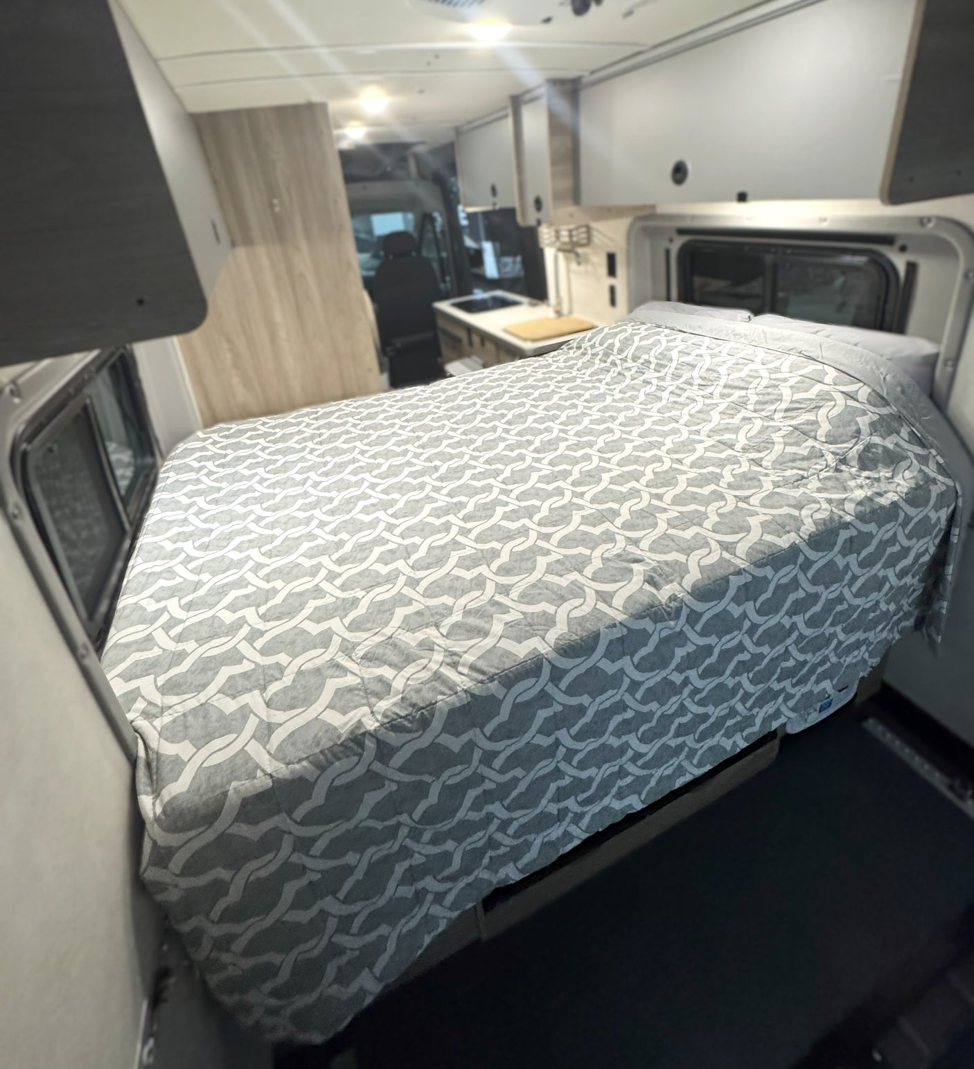 Cozy and compact RV rental interior featuring a large bed with a gray patterned quilt, and a kitchen area in the background.