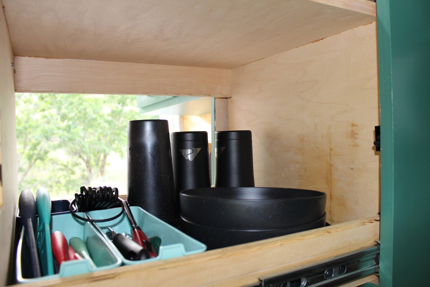 Efficient RV rental cabinet storage with black cups, bowls, and organized teal kitchen tools, showing a nature view outside. — photo 3