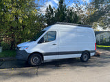 White Mercedes-Benz Sprinter van with roof rack, a perfect camper van for RV rental, parked on asphalt with trees.