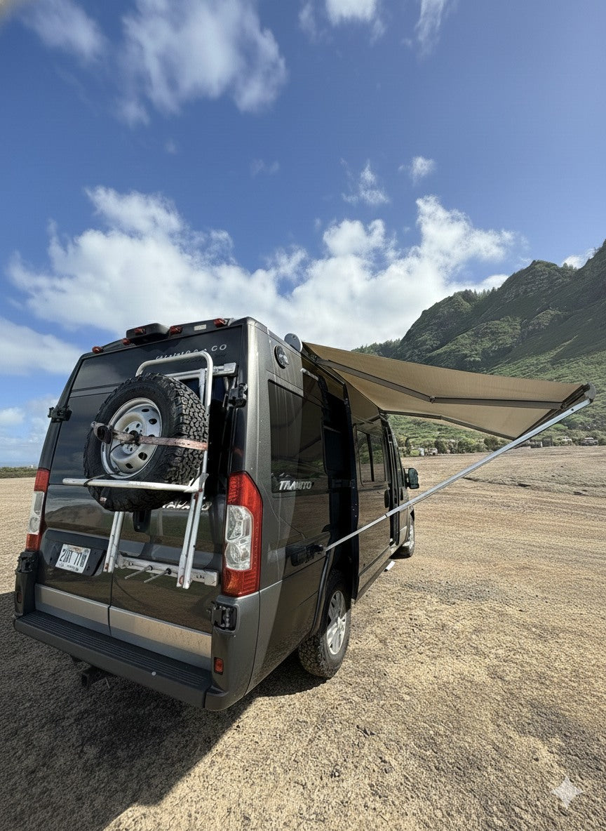 Dark gray RV rental, a camper van with an awning extended, parked on sandy ground with green mountains & blue sky.