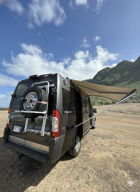 Dark gray RV rental, a camper van with an awning extended, parked on sandy ground with green mountains & blue sky.