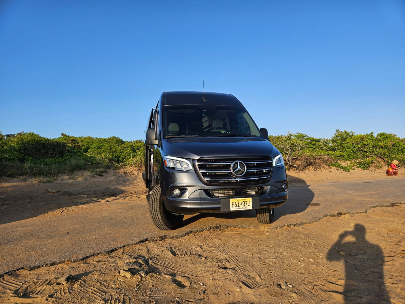 Front view of a dark gray Mercedes-Benz Sprinter van RV rental on a sandy road by green bushes under a clear sky.