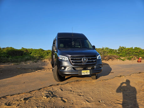 Front view of a dark gray Mercedes-Benz Sprinter van RV rental on a sandy road by green bushes under a clear sky.