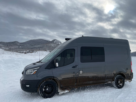 Muddy grey RV rental van in snowy mountains under cloudy sky, ready for winter adventure.