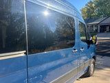 Blue Sprinter van for RV rental, parked under sun, reflecting trees and a house on its windows.
