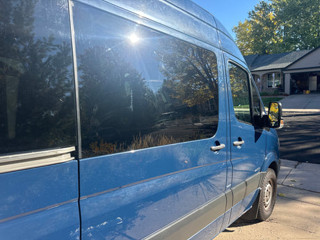 Blue Sprinter van for RV rental, parked under sun, reflecting trees and a house on its windows.