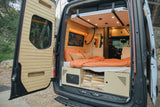 Cozy camper van interior of an RV rental, featuring a bed, storage, and custom wooden walls, open to nature.