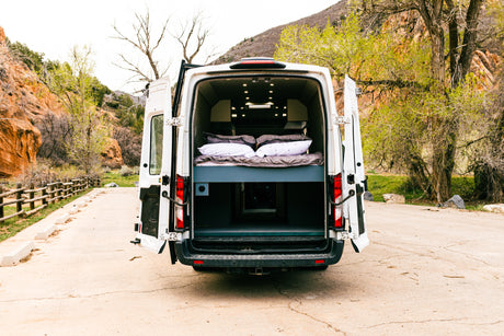 Rear view of a white camper van with doors open, revealing a comfy bed and storage.