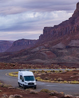 White rv rental campervan parked on a winding desert road, surrounded by majestic red cliffs and a dramatic sky.