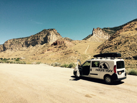 Explore the wild! White Nature Vans camper van, an RV rental option, parked on a dirt road in arid mountains under blue sky.