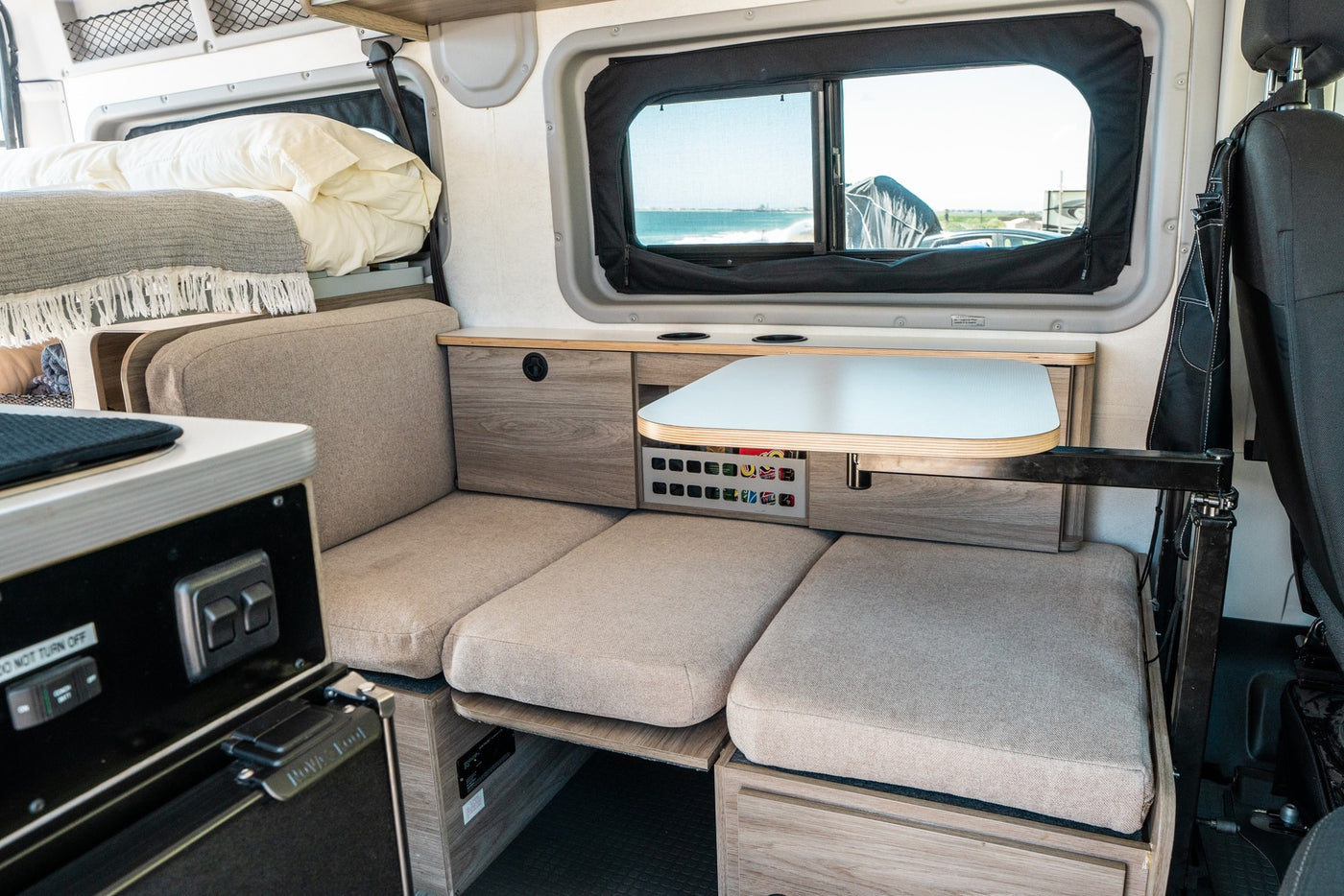 Inside a modern rv rental: plush bed, adaptable bench seating, and pull-out table with a bright ocean window view. — photo 14