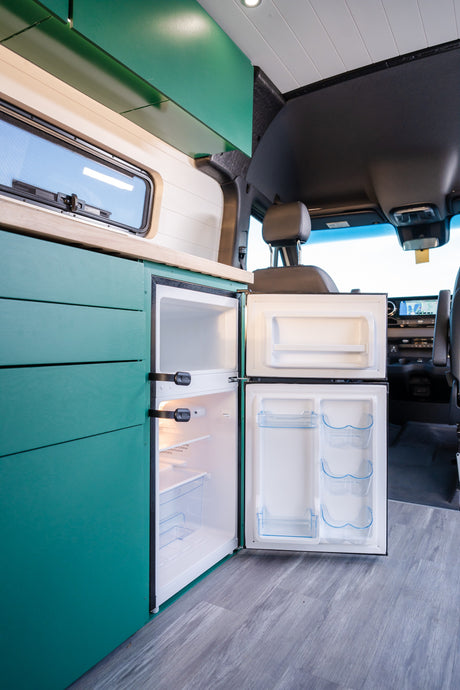 Inside this RV rental: an open mini-fridge, sleek teal cabinetry, wood counter, and window in a modern camper van.