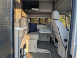Inside a modern rv rental: a dining area with bench seating, small table, and overhead TV. A fire extinguisher is visible.