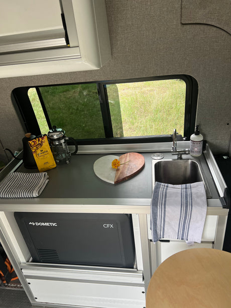 Compact RV rental kitchen with sink, Dometic fridge, and coffee setup. Enjoying van life with a view of green fields.