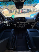 Inside a Mercedes-Benz RV rental: the front cabin showing dashboard, steering wheel, and black leather seats, facing a residential street.