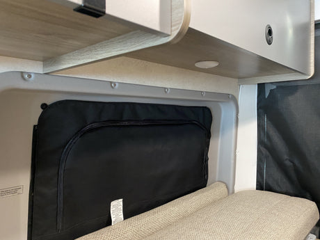Compact RV rental interior: overhead storage, black fabric storage/cover, and a cozy seating area with visible warning labels.