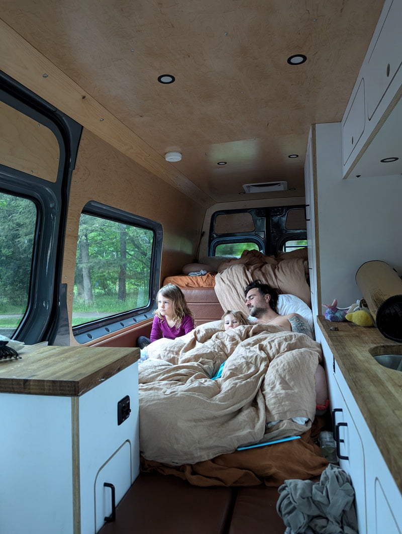 Inside a comfortable RV rental: A family, dad and two children, are snuggled in bed, gazing out the van's window at the surrounding trees. — photo 15