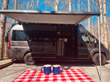 Enjoying coffee by our RV rental with the awning out. A grey camper van is parked under a bright blue sky among bare trees.
