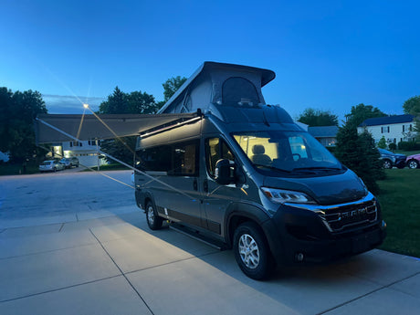 A sleek dark grey pop-top campervan rv rental with its awning extended and lights on, parked in a driveway at dusk.