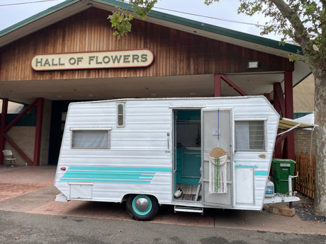 Vintage white & teal RV rental outside "Hall of Flowers". Open door reveals retro interior design.