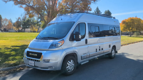 A silver Ram camper van, a great RV rental option, parked on a street beside a vibrant autumn park.