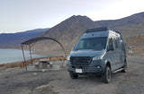 Gray Mercedes Sprinter rv rental on a scenic lake shore with mountains and a picnic table. Ideal for adventure.