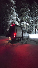 An RV rental shines in a snowy forest at night, its red taillights illuminating fresh tracks for winter van camping adventure.