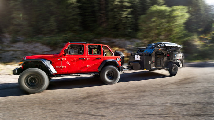Red Jeep Gladiator towing a rugged black rv rental for off-road camping, in motion on a scenic road.