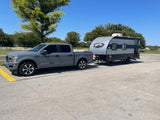 A grey truck tows a compact travel trailer on asphalt under a clear sky. Ideal for your next RV rental and camping adventure!