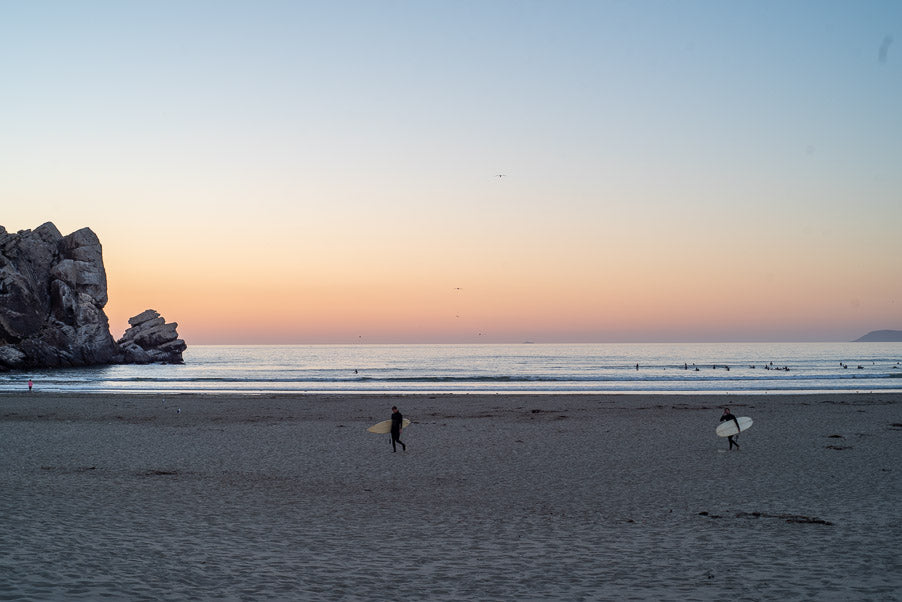 Surfers on a sandy beach with rock formations at sunset. Imagine a relaxing RV rental trip ending here.