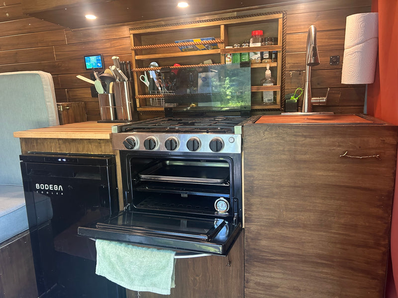 Inside an RV rental, a compact kitchen features a gas stove/oven, sink, and Bodega cooler amidst rustic wooden finishes. — photo 7